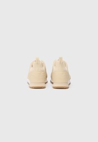Lys beige treningssko med teksturert overdel, minimalistisk design, pull-tabs og såle i hvit og tan. Har preget logo på hælen.