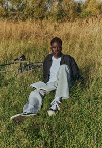 Personne assise sur l'herbe, portant une veste noire sur un t-shirt blanc, un jean en denim clair et des baskets noires, tenant un livre ouvert. Vélo à proximité.