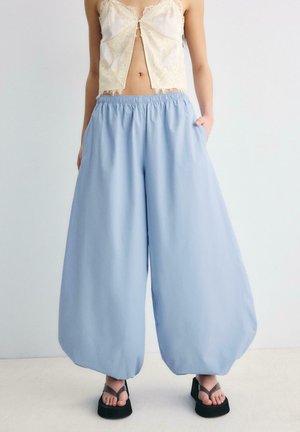 Frau trägt lockere hellblaue Hose, cremefarbenes Spitzenoberteil und schwarze Plateau-Sandalen, steht mit den Händen in den Taschen.