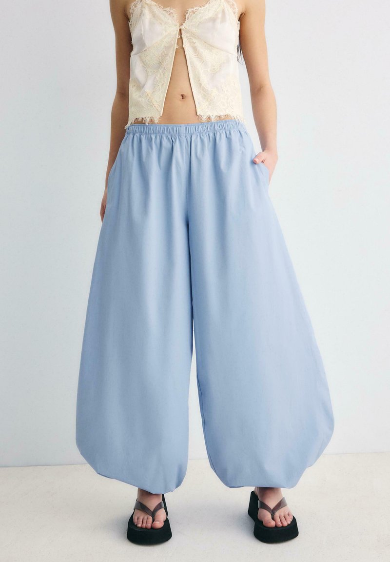 Femme portant un pantalon ample bleu clair, un haut en dentelle crème et des sandales à plateforme noires, debout les mains dans les poches.