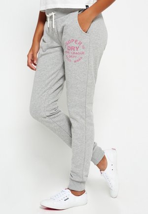 Person trägt hellgraue Jogginghose mit pinkem "Super Dry"-Logo am Oberschenkel, weiße Sneakers und weißen Kordelzugbund.