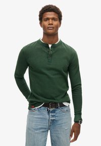 Camisa henley de manga larga en verde oscuro con un acabado texturizado, tapeta de tres botones y puños acanalados, combinada con vaqueros azul claro y un cinturón negro.