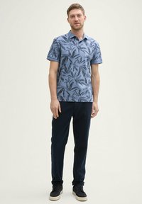 Polo shirt bleu à manches courtes avec un motif de feuilles noires, associé à un pantalon bleu foncé et des baskets noires avec des semelles blanches.