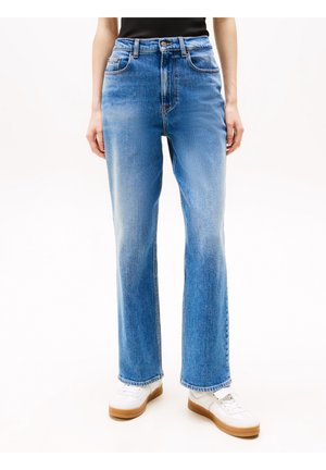 Personne portant un jean bleu clair coupe droite, des baskets blanches avec semelles en gomme et un haut noir, debout devant un fond blanc.