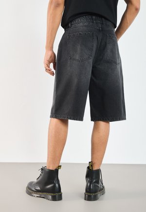 Osoba ubrana w czarne szorty z denimu sięgające kolan, czarny T-shirt oraz czarne sznurowane buty z żółtym przeszyciem, stojąca na szarej podłodze.
