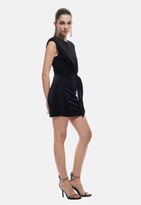 Robe noire ajustée avec un détail avant torsadé et des manches courtes. Texture lisse avec un léger éclat. Associée à des talons hauts noirs à lanières.