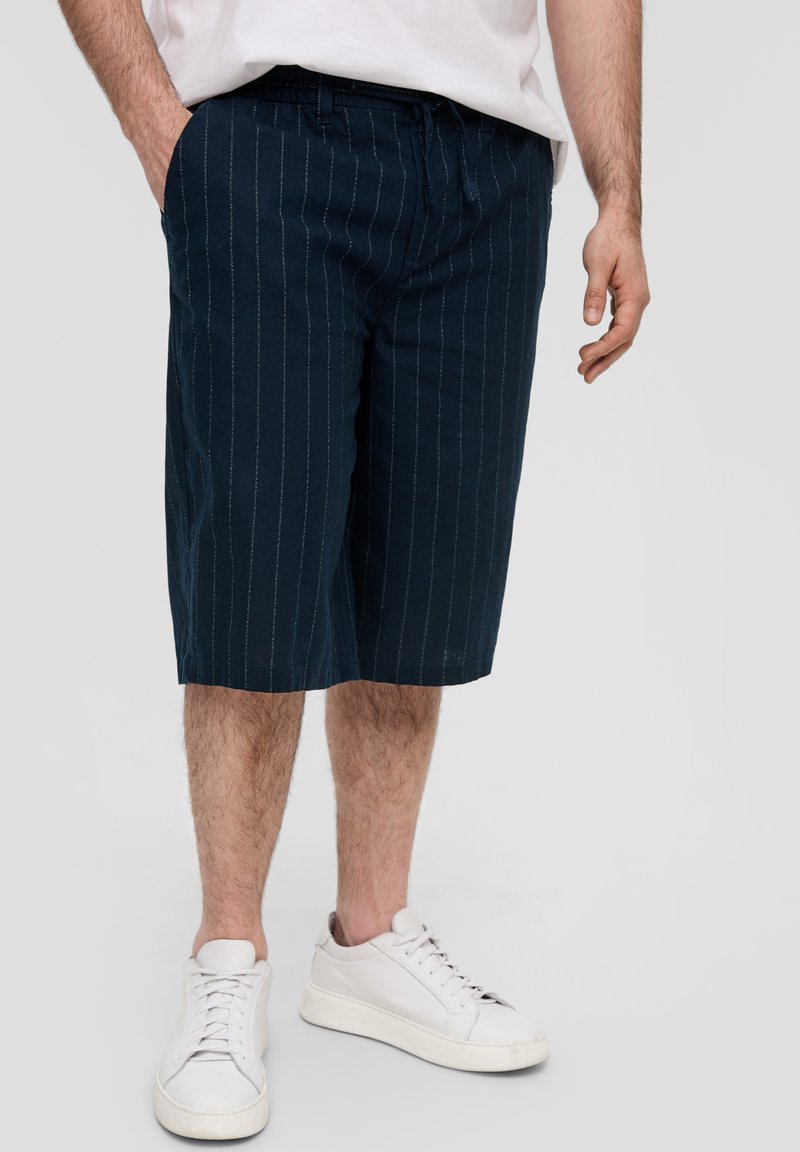 Shorts marins avec des rayures verticales blanches, fabriqués en tissu doux. Comprend une taille élastique et des poches latérales. Associés à des baskets blanches.