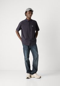 Camisa de mangas curtas, azul marinho, com colarinho abotoado e logótipos no peito, combinada com jeans azuis claros e ténis bege. Boné cinza.