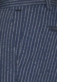 Pruhovaná modrá látka s tenkými bílými liniemi a texturovaným povrchem. Obsahuje kapsu a hladký detail pasu.