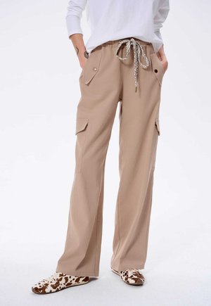 Beige Cargo-Hosen mit Kordelzugbund, zwei Seitentaschen und einer lockeren Passform. Kombiniert mit gemusterten Slippern.