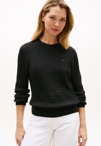 Schwarzer, strukturierter Strickpullover mit rundem Ausschnitt und gerippten Bündchen. Mit einem kleinen Logo auf der Brust. Kombiniert mit weißen Hosen.