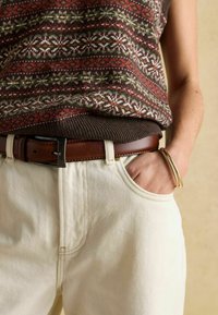 Oseba v puloverju s vzorcem, zategnjenem v kremne hlače z rjavim usnjenim pasom, z roko v žepu in dvema zlatima zapestnicama na zapestju.