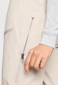 Beežist kangast välirõivaste kombinesoon koos küljerihmaga lukuga taskuga ja halli pika varrukaga sisemisest särgist detailiga. Ühtlane tekstuur, puhtad jooned, minimalistlik disain.