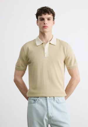 Jeune homme portant un polo beige à manches courtes avec un col blanc et un jean bleu clair, debout les mains derrière le dos.