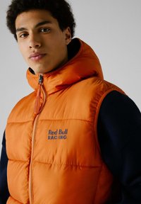 Narančasti puffasti prsluk s kapuljačom, s patentnim zatvaračem i logotipom "Red Bull Racing" u tamnoplavoj boji. Nositi preko tamnoplave veste.