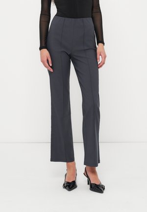 Vrouw staat tegen een effen achtergrond gekleed in een donkergrijze high-waist broek, zwarte top met transparante mouwen en zwarte hakken.