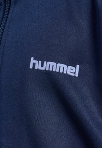 Marine-blaue Sportbekleidungsstoff mit glatter Textur, mit einem auffällig gedruckten weißen "hummel"-Logo. Der Reißverschluss ist seitlich sichtbar.