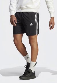 Μαύρα αθλητικά σορτς με λευκή τριπλή ρίγα, κατασκευασμένα από ελαφρύ υλικό. Φορεμένα με λευκές κάλτσες και μαύρα αθλητικά παπούτσια.