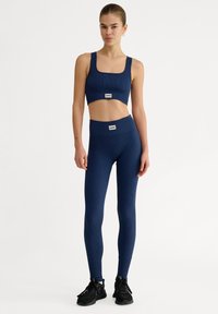Marinblå sport-bh med ribbad design och matchande högmidjade leggings, med en kort topp och logodetaljer, stylade med svarta träningsskor.