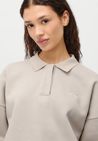 Lys beige polo-skjorte med krage, med en brodert "LeGer" logo i hvitt på venstre side. Myk, teksturert stoff.