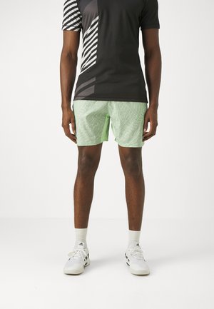 Tenue sportive comprenant un t-shirt noir à manches courtes avec des rayures blanches diagonales et un short à motifs verts, associé à des chaussures de sport blanches.
