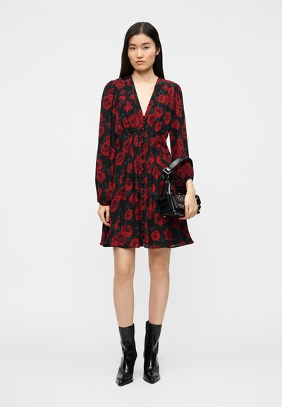 Μαύρο φόρεμα με βαθύ V λαιμόκοψη, μακριά μανίκια και σχέδιο από κόκκινο τριαντάφυλλο. Φορεμένο με μαύρα μποτάκια αστραγάλου και κρατώντας μια μικρή μαύρη τσάντα.