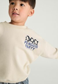 Sweatshirt crème avec un col rond, affichant un graphique de pingouin cartoon et le texte "PENGUIN PARTY" en bleu. Tissu doux et texturé.