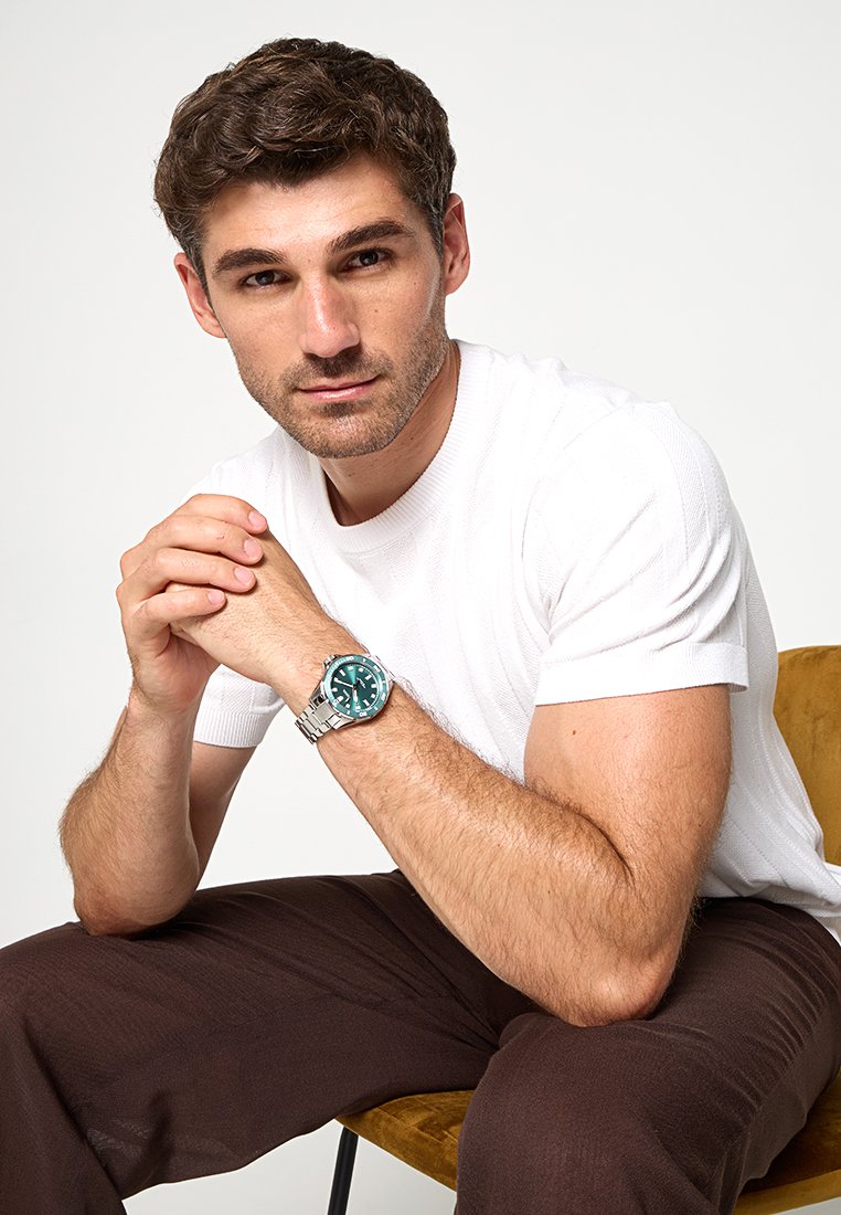 Silberne Uhr mit türkisfarbenem Ziffernblatt am Handgelenk, getragen von einem Mann in einem weißen strukturierten T-Shirt und braunen Hosen, der auf einem gelben Stuhl sitzt.
