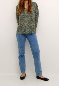 Blouse verte à motif cachemire avec un col en V et des manches longues, associée à un jean bleu clair coupe droite et des ballerines noires.