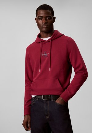 Άνδρας που φοράει κόκκινη μπλούζα Calvin Klein με μπροστινή τσέπη και σκούρο τζιν, στέκεται μπροστά σε απλό ανοιχτό γκρι φόντο.
