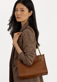 Καφέ δερμάτινη τσάντα χειρός με υφή, αλυσίδα σε χρυσόχρωμο τόνο και ανάγλυφο λογότυπο. Διαθέτει ορθογώνιο σχήμα και ελάχιστο υλικό. 