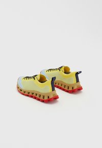 Sport shoes featuring a yellow knitted upper, blue accents, black laces, and a thick tan midsole with red rubber sole, designed for cushioning.