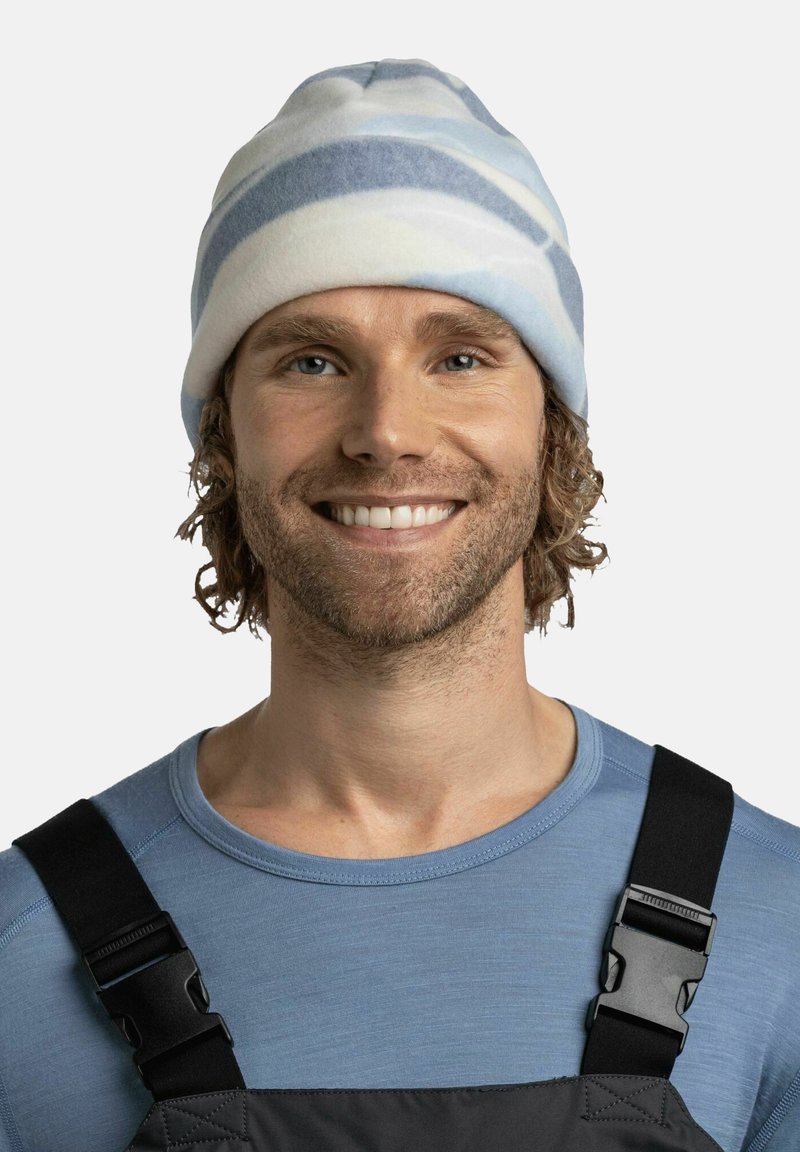 Gorro de rayas azul claro y blanco, hecho de tela suave, con un ajuste ceñido. El hombre lo lleva con una camiseta de manga larga azul y un overol oscuro.