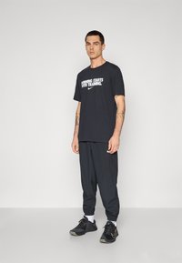 Juodas medvilninis marškinėlis su balta tekstu "LAIMĖ PRASIDEDA NUO TRENIRUOČIŲ" ir Nike logotipu, derinamas su juodais siaurais keliais ir juodais sportbačiais.