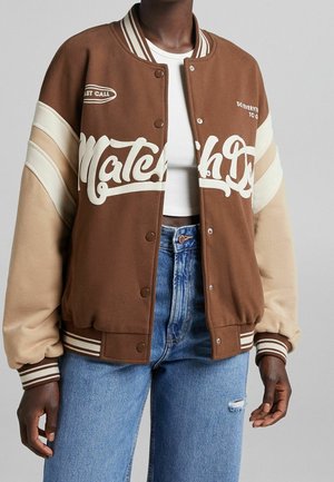 Persona que lleva una chaqueta varsity marrón y beige con letras blancas sobre un top corto blanco y vaqueros azules de cintura alta con un desgarrón en el muslo.