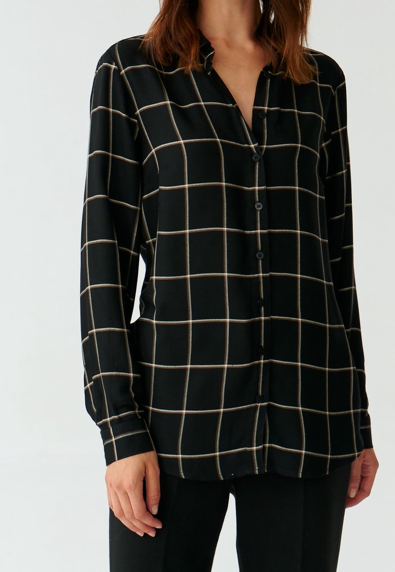 Femme portant une chemise noire à boutons avec des carreaux blancs et beiges en motif quadrillé, associée à un pantalon noir, vue du cou jusqu'aux hanches.