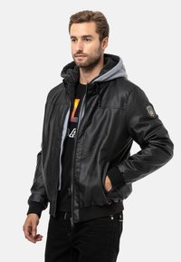 Schwarze Lederjacke mit grauem Hoodie-Futter, Rippbündchen, Frontreißverschluss, zwei seitlichen Taschen und einem Abzeichen-Detail am Ärmel.