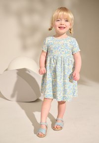 Blommönstrad klänning i pastellfärger med korta ärmar, markerad midja och vågiga kanter. Matchas med ljusblå sandaler.