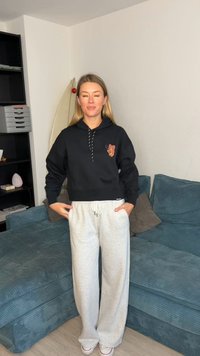 Črna kapucar s krajšim krojem in motivom metulja, kombinirana s svetlo sivimi trenirkami. Kapucar ima vrvico za zategovanje in rebraste manšete.