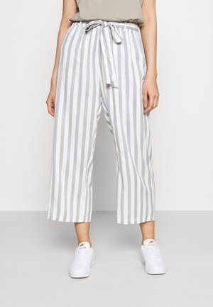 Persoon draagt witte en lichtgrijze wijde broek met verticale strepen, een beige top en witte sneakers.