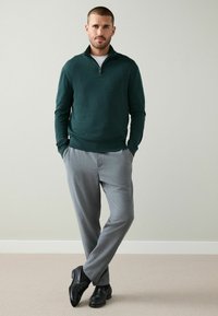 Mörkgrön zip-up-tröja med långa ärmar, kombinerad med ljusgrå byxor och svarta finskor, stående på ett ljust heltäckningsmatta golv.