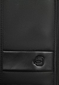 Tela negra texturizada con un acento liso de cuero. Presenta un logo sutil y una cremallera a lo largo del borde para el cierre.