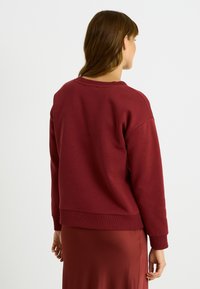 Sudadera burdeos con cuello redondo, puños y dobladillo acanalados. Textura suave, corte relajado y hombros caídos; usada con una falda a juego.