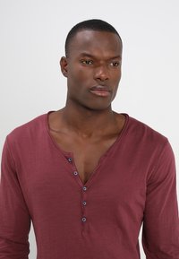 Weinrote, langärmlige Henley-Shirt mit V-Ausschnitt, vier Knöpfen und geripptem Kragen. Weicher Stoff mit leichter Textur.
