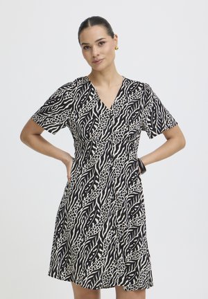 Kobieta z włosami związać do tyłu, ubrana w krótką sukienkę w czarno-biały zwierzęcy wzór, stoi z rękami na biodrach na tle jednolitym.