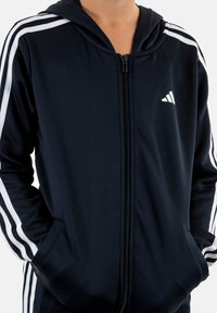 Felpa con zip di colore blu navy con strisce bianche sulle maniche. Realizzata in tessuto morbido, presenta una zip frontale e tasche laterali. Logo sulla parte superiore sinistra del petto.