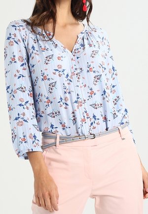 Frau trägt eine hellblaue, geblümte Bluse mit dreiviertel Ärmeln, in helle rosa Hose mit schmalem Gürtel gesteckt.