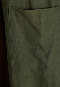 Tissu en lin vert olive avec une poche visible, présentant une texture lisse et un design simple et fonctionnel. Détail de couture minimaliste présent.