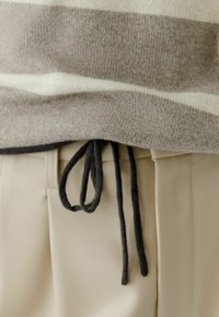 Beige broek met een zwarte trekkoord in een strik geknoopt, gecombineerd met een beige gebreide trui met een witte horizontale streep.