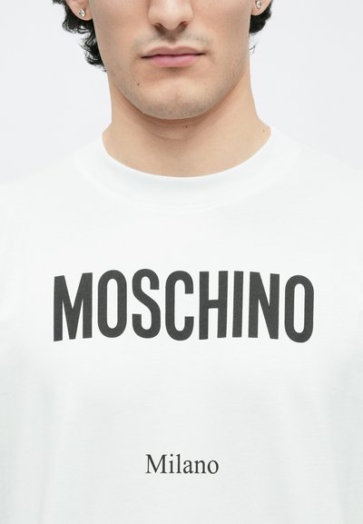 Biały bawełniany t-shirt z wyraźnym czarnym napisem "MOSCHINO" nad mniejszym napisem "Milano", czystymi liniami i okrągłym dekoltem.